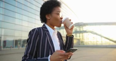 4 bin. Çekici Afrikalı Amerikalı kadın telefonundan bir şey okurken gülümsüyor ve akşam güneşinin altında modern binadan önce kahve içip yürüyor.