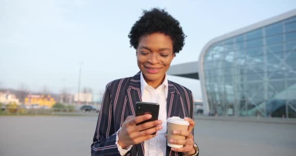 4k. Zeitlupe. Positive Emotionen. Afroamerikanische Geschäftsfrau im stylischen Strip-Anzug spaziert mit einer Tasse Kaffee to go und liest etwas in ihrem Smartphone — Stockvideo