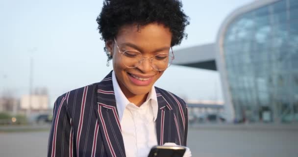 4 bin. Ağır çekim. Olumlu duygular. Şık giyinmiş Afro-Amerikalı iş kadını elinde bir fincan kahveyle yürüyüp akıllı telefonundan bir şeyler okuyor. — Stok video