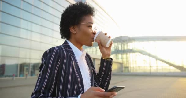 4k 。慢动作迷人的非洲裔美国女人一边看手机里的内容，一边在夕阳西下的现代建筑前喝咖啡，一边笑着 — 图库视频影像