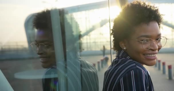 4k. En cámara lenta. Emociones positivas. Encantadora mujer afroamericana en gafas y elegante traje despojado sonríe de pie ante una pared de vidrio de edificio moderno — Vídeo de stock