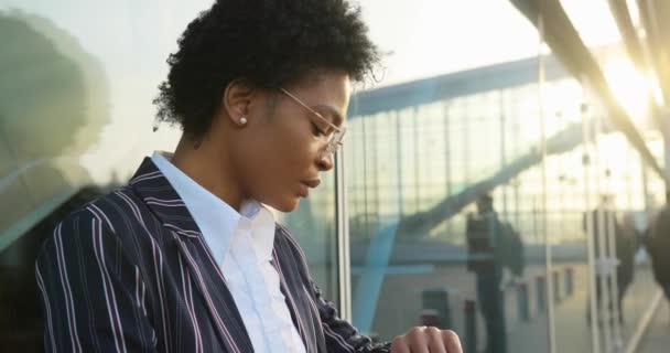 4k. Αργή κίνηση. Αφρο-Αμερικανίδα επιχειρηματίας με κομψό ριγέ κοστούμι κοιτάζει το ρολόι της και στέκεται ανήσυχος μπροστά σε ένα σύγχρονο κτίριο — Αρχείο Βίντεο