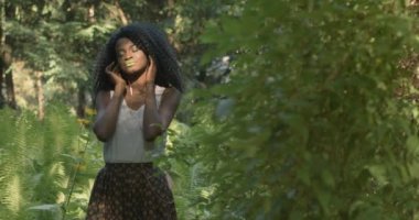 4 bin. Hızlı ve yavaş çekim. Teknoloji kullanımı. Yeşil gözlü ve dudaklı çekici Afrikalı Amerikalı kadın yeşil egzotik ormanda duran kulaklıklarıyla müzik dinliyor.