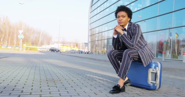 4k. Мандрівка, цифровий. Чарівна афро-американська жінка в сучасному смугастому костюмі сидить біля скляної стіни на її дорожній сумці. Безробітна жінка або студент. — стокове відео