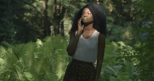 4k. Veloce e lento. Utilizzo della tecnologia. Attraente donna afroamericana con occhi verdi e labbra parla sullo smartphone e ride in piedi nella foresta esotica del mattino — Video Stock