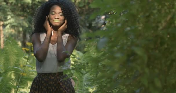 4k. Γρήγορη και αργή κίνηση. Χρήση τεχνολογίας. Ελκυστική Αφροαμερικανή γυναίκα με πράσινα μάτια και χείλη ακούει τη μουσική στα ακουστικά της να στέκεται στο πράσινο εξωτικό δάσος — Αρχείο Βίντεο