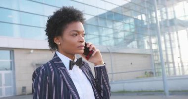 4 bin. Seyahat, digtal, iş. Şık giyinmiş çekici Afrikalı Amerikalı kadın modern cam bir binanın önünde yürürken telefonla konuşuyor.