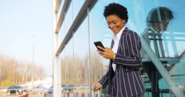 4 bin. Seyahat et, Dijital. Modern giyinmiş Afro-Amerikalı çekici kadın cam duvarın yanında duruyor ve telefonunda bir şeyler okuyor. İş kadını ya da öğrenci