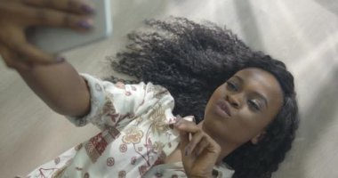Genç ve güzel Afrikalı kızın portresinin üstünde sevimli bir gülümseme ve doğal bir makyajla yerde yatarken cep telefonuyla selfie çekiyor.