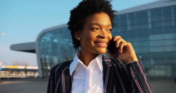 4k. Viajes, digestivos, negocios. Atractiva mujer afroamericana en traje despojado con estilo habla por teléfono caminando ante un moderno edificio de vidrio — Vídeo de stock