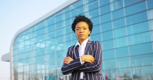 4k. Reizen, zaken doen, levensstijl. Aantrekkelijke Afro-Amerikaanse vrouw in gestript pak kruist haar armen op de borst staande voor een modern glazen gebouw in een zonnige dag — Stockvideo