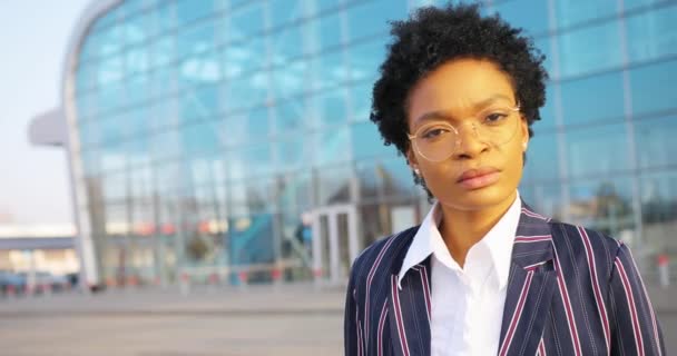 4k. Reizen, zaken doen, levensstijl. Aantrekkelijke Afro-Amerikaanse vrouw in gestript pak kruist haar armen op de borst staande voor een modern glazen gebouw in een zonnige dag — Stockvideo
