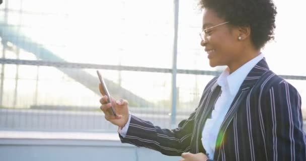 4k. Negócios, estilo de vida, viagens. Emoções positivas. Mulher afro-americana atraente em terno despojado elegante leva uma selfie em seu telefone de pé fora — Vídeo de Stock