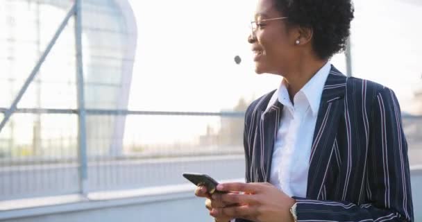 4k. Reizen, zaken doen, levensstijl. Aantrekkelijke Afro-Amerikaanse vrouw in gestript pak ziet er gelukkig uit wandelen voor een modern glazen gebouw in een zonnige dag en iets lezen in haar telefoon — Stockvideo