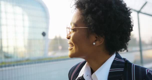 4k. Viaggi, affari, stile di vita. Attraente donna afroamericana in abito spogliato sembra felice di camminare davanti a un moderno edificio di vetro in una giornata di sole — Video Stock