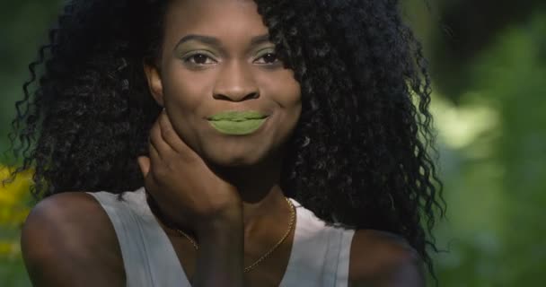 4k. Movimento rápido e lento. Mulher afro-americana atraente com olhos e lábios verdes olha diretamente na câmera e sorri em pé na floresta exótica — Vídeo de Stock