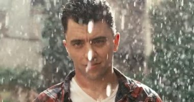 Odak değiştiriyorum. Gündelik giysiler içindeki çekici sakin adam yağmurda kameraya bakıyor. 4k görüntü.