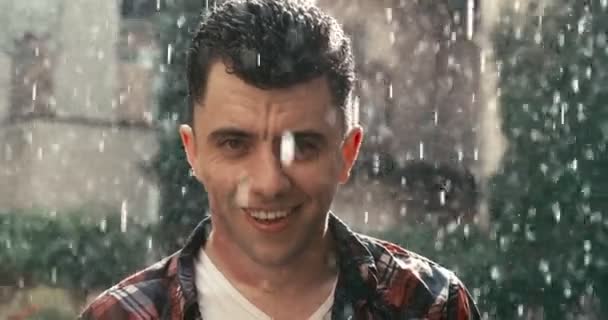 Närbild porträtt av den unga stiliga mannen med mörkt hår och härligt leende njuter av regnet under den soliga dagen. 4k-bilder. — Stockvideo