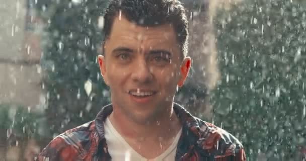 Positives Porträt des attraktiven Typen, der lächelt und in die Kamera schaut, während er den schönen Regen während des sonnigen Tages genießt. 4k Filmmaterial. — Stockvideo