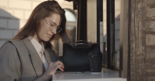 チャーミングな笑顔で眼鏡の素晴らしい若いビジネスマンの女性がコーヒーブレイクをしています。彼女は携帯電話でメールとブラウジングをしている。4k. — ストック動画