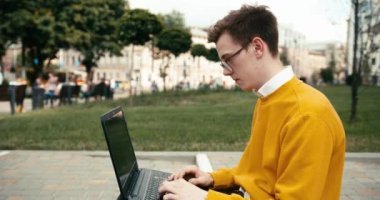 Gözlüklü, dışarıda dizüstü bilgisayarda çalışan genç sarışın adamın yan portresi. 4k görüntü.