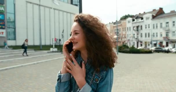 Attraktiv glad ung flicka med charmigt leende och hängslen talar på mobilen när du går längs gatan. — Stockvideo
