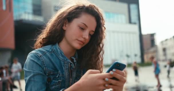 Güzel gülüşü ve diş telleri olan mutlu sevimli kız dışarıda cep telefonuyla mesajlaşıp sohbet ediyor. Duygusal Portre. — Stok video