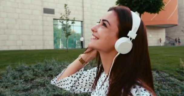 Happy urocza dziewczyna z ciemnymi włosami i piękny uśmiech jest korzystających z muzyki w słuchawkach podczas spaceru wzdłuż ulicy. — Wideo stockowe