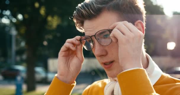 Retrato lateral del guapo adolescente con el pelo rubio poniéndose gafas modernas al aire libre. Imágenes de 4k. — Vídeos de Stock