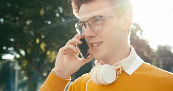 Young handsome teenager with blond hair in eyeglasses is happily talking on the mobile phone in the street. 4k footage. — Stock Video