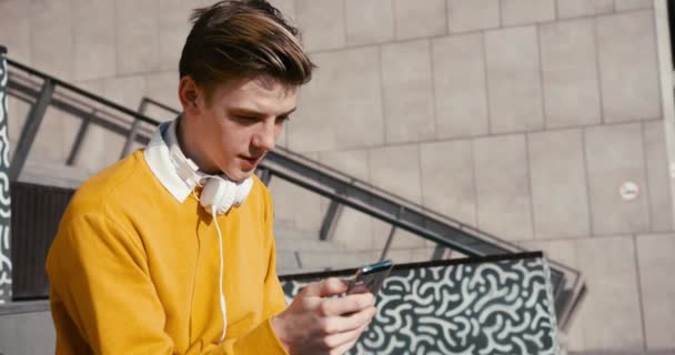 Jovem hipster atraente com cabelo loiro em suéter amarelo está conversando e navegando no telefone celular. Filmagem 4k. — Vídeo de Stock