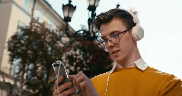 Молодь і технології. Привабливий молодий хлопець зі світлим волоссям, окулярами та веснянками спілкується на мобільному телефоні та слухає музику через навушники на відкритому повітрі . — стокове відео