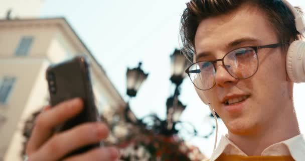 ヘッドフォンの屋外を介して音楽を聴きながらチャットのためのスマートフォンを使用して眼鏡の魅力的な男の肖像画をアップ. — ストック動画