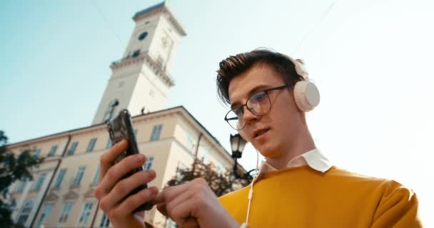 Портрет вгору. Люди і технології. Привабливий хлопець в окулярах серфінг мережі за допомогою свого смартфона і прослуховування музики на відкритих навушниках. 4k кадри . — стокове відео
