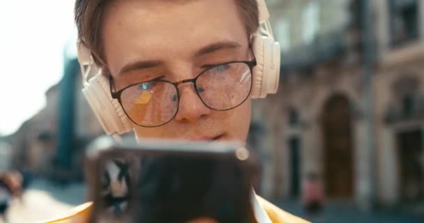 Передній крупним планом привабливий усміхнений хіпстер в окулярах тримає смартфон і насолоджується музикою в білих навушниках на відкритому повітрі. 4k кадри . — стокове відео