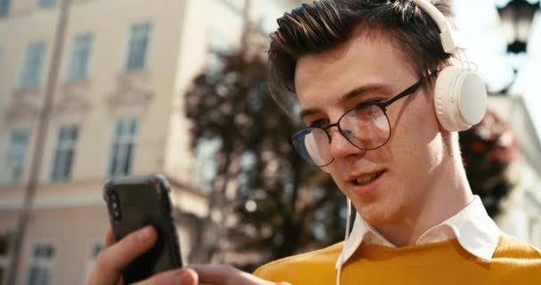 サイドポートレート。眼鏡の魅力的な若いブロンドの男は、ヘッドフォンの屋外で音楽を楽しみながら、スマートフォンでチャット、ブラウジング、テキストメッセージ. — ストック動画