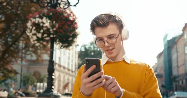 眼鏡の中のハンサムな若いヒップスターは、スマートフォンでチャットや閲覧しています。彼は電話で音楽を楽しんでいる。サニーシティセンターの場所。4k映像. — ストック動画