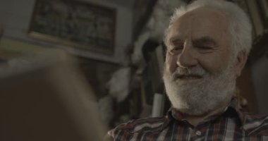 Gri sakallı yaşlı gülümseyen sanatçı atölyesinde bir resim çiziyor. 4k.