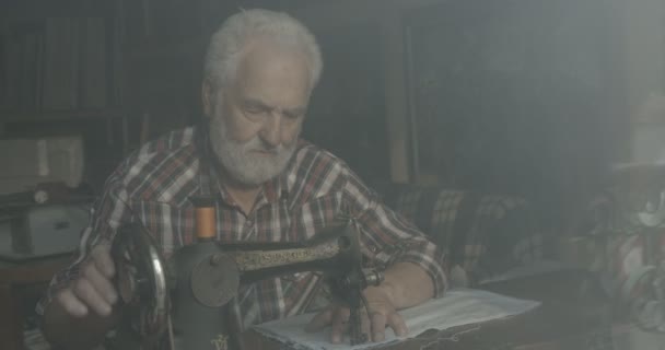 Leeftijd geïnspireerde oude man. Hij naait op een oude handgenaaide machine in zijn werkplaats. Portret. 4k. — Stockvideo
