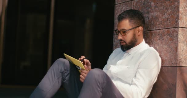 Ο αφρο-αμερικανός γραφίστας στα γυαλιά εργάζεται πάνω στο ψηφιακό σχεδιαστικό tablet ενώ κάθεται σε εξωτερικό χώρο. 4k πλάνα. — Αρχείο Βίντεο