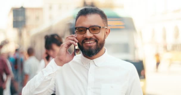 Portret van de blije glimlachende Afrikaanse man in bril emotioneel pratend op de mobiele telefoon in de straat. — Stockvideo