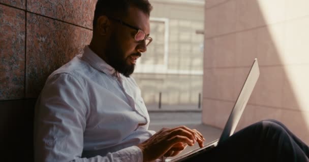 O triste homem de negócios africano cansado em óculos está digitando no laptop enquanto está sentado na rua. — Vídeo de Stock