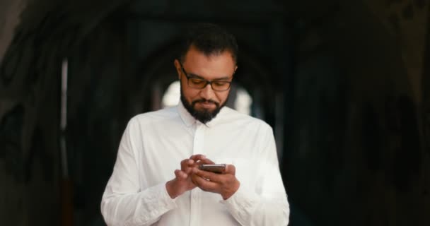Retrato do homem africano sorridente alegre em óculos está enviando mensagens, conversando e navegando no telefone celular no fundo preto ao ar livre. — Vídeo de Stock