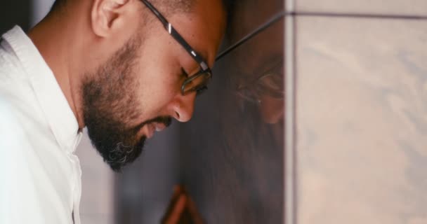 O belo homem africano deprimido de óculos está inclinado na parede e a bater-lhe com o punho. Retrato de close-up lateral. — Vídeo de Stock