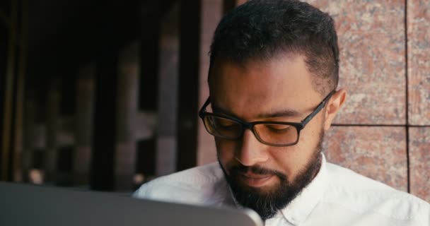 Крупним планом портрет засмученого африканського бізнесмена в окулярах, що працюють, спілкуються і переглядають ноутбук на відкритому повітрі . — стокове відео