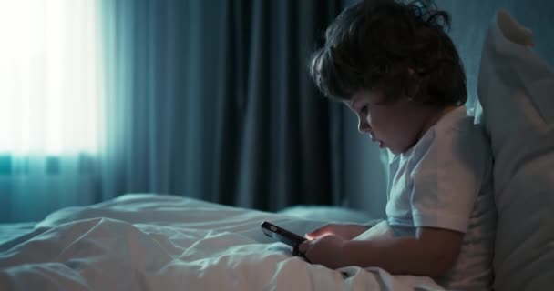 Jonge mooie jongen zit in wit bed en doorzoek iets aan de telefoon. 4K-video — Stockvideo