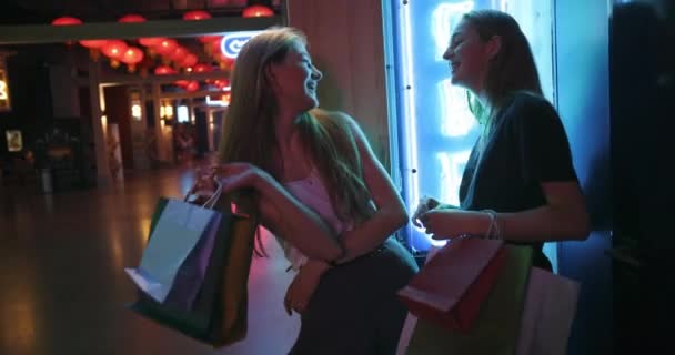 Zwei junge hübsche Damen, die nach erfolgreichem Einkauf spazieren gehen, lächeln und halten Farbpakete in den Händen. Stilvolle Mädchen spazieren durch das Einkaufszentrum und haben Spaß. Ein spannendes Gespräch zwischen