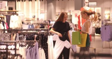 Güzel kızlar butikte alışveriş yaparken kıyafet seçiyor, konuşuyor ve gülümsüyor. Alışveriş yapan iki genç kadının portresi. 4K video