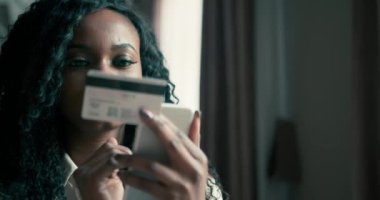 Kıvırcık saçlı güzel Afrikalı Amerikalı kadın cep telefonu ve kredi kartıyla internetten bir şeyler alıyor. Neşeli bir esmer tarafından internetten alışveriş yapmak. 4K video