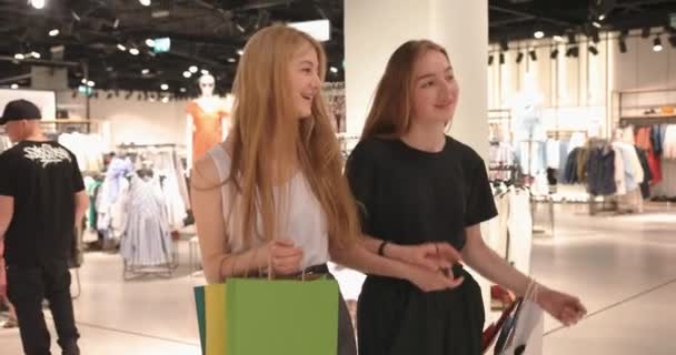 Vackra tjejer väljer kläder, pratar och ler medan de shoppar i butiken. Porträtt av två vänner som går med sina inköp i klädaffären och shoppar. 4K-video — Stockvideo